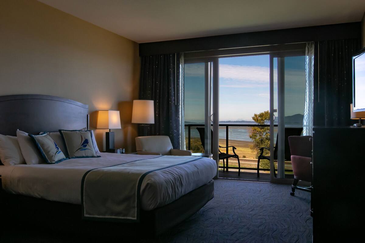 Habitación de hotel con una cama grande, dos lámparas de noche, un escritorio, una silla y un balcón con vistas a la playa y al océano.