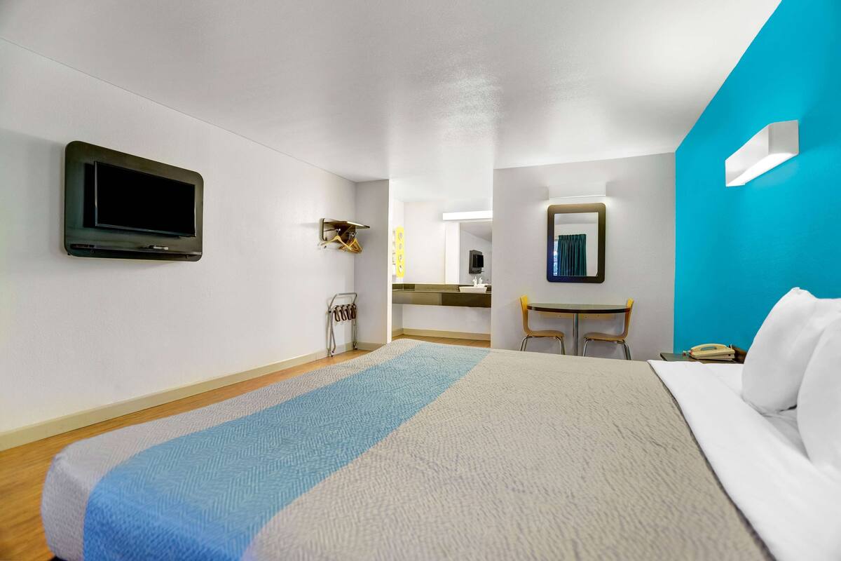 A hotel room with a bed, a desk, a chair, a TV, and a bathroom visible in the background.