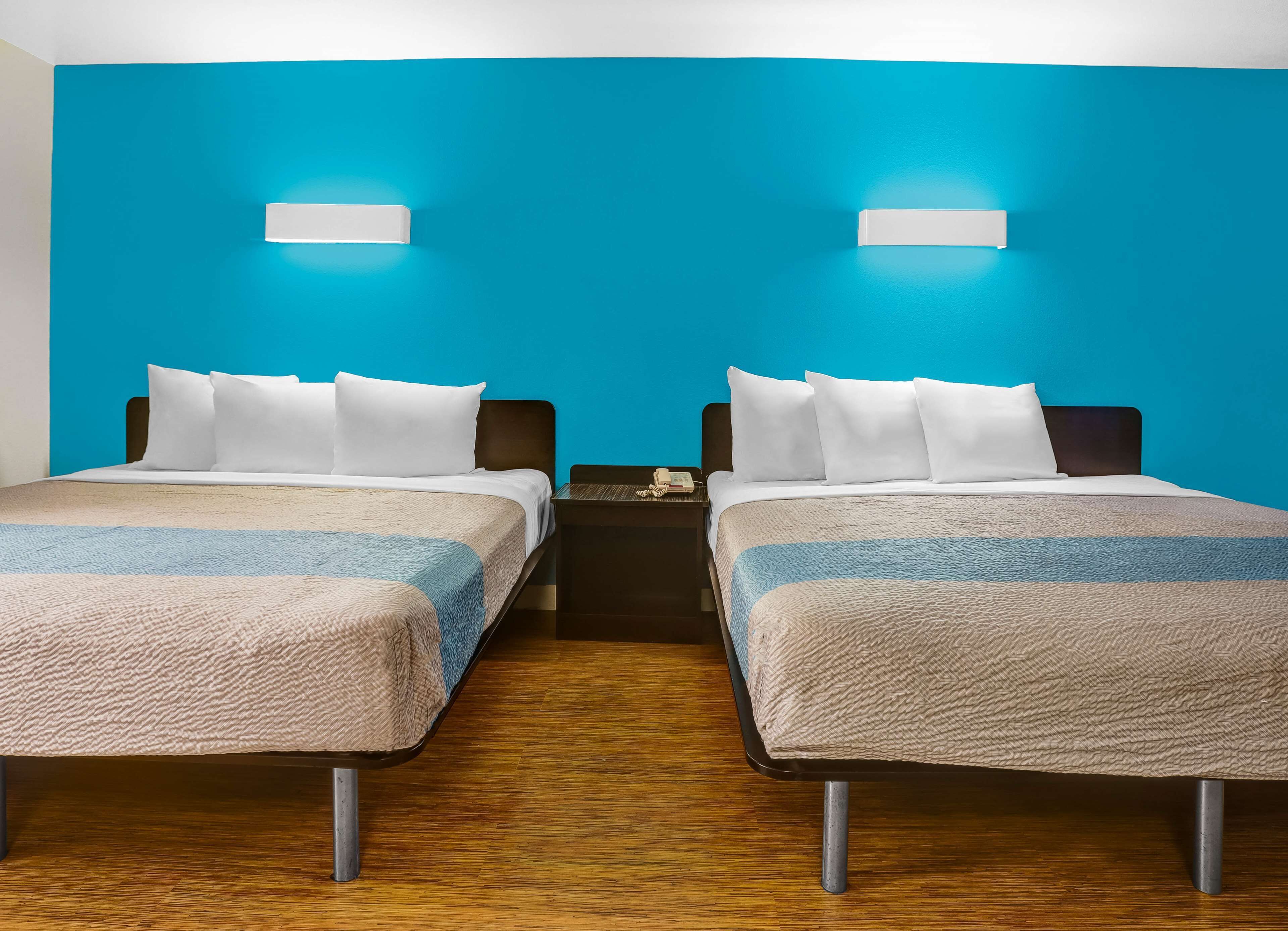 A hotel room with two beds, each with white bedding and a blue and beige striped blanket. There are two bedside tables with lamps and a phone. The walls are painted blue, and the floor is wooden.
