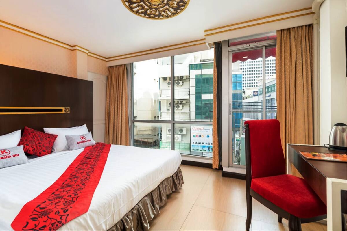 A hotel room with a large bed, a red chair, a desk, and a view of the city through a large window.