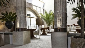 Un amplio espacio interior con piso de piedra, sillas blancas y grandes macetas con plantas.