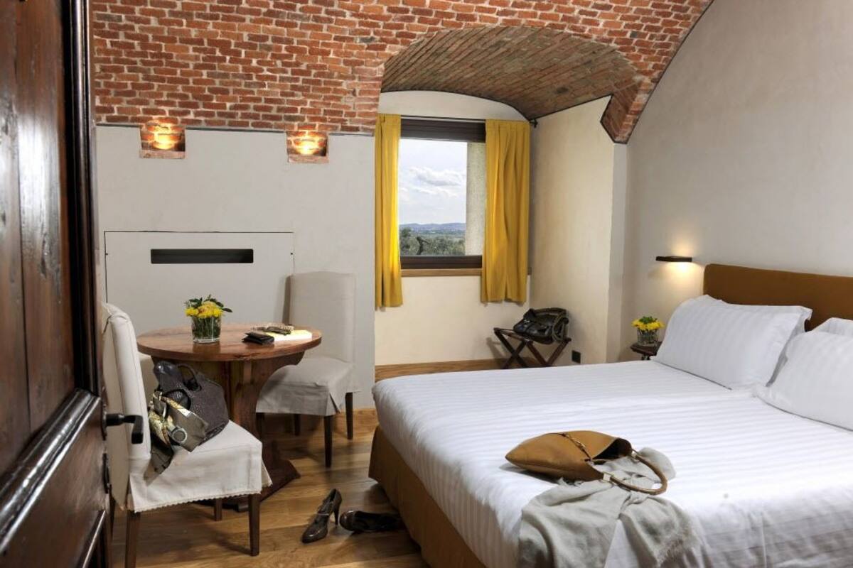 A hotel room with a brick ceiling, a bed with white bedding, a small round table with a vase of flowers, a chair, and a window with yellow curtains.