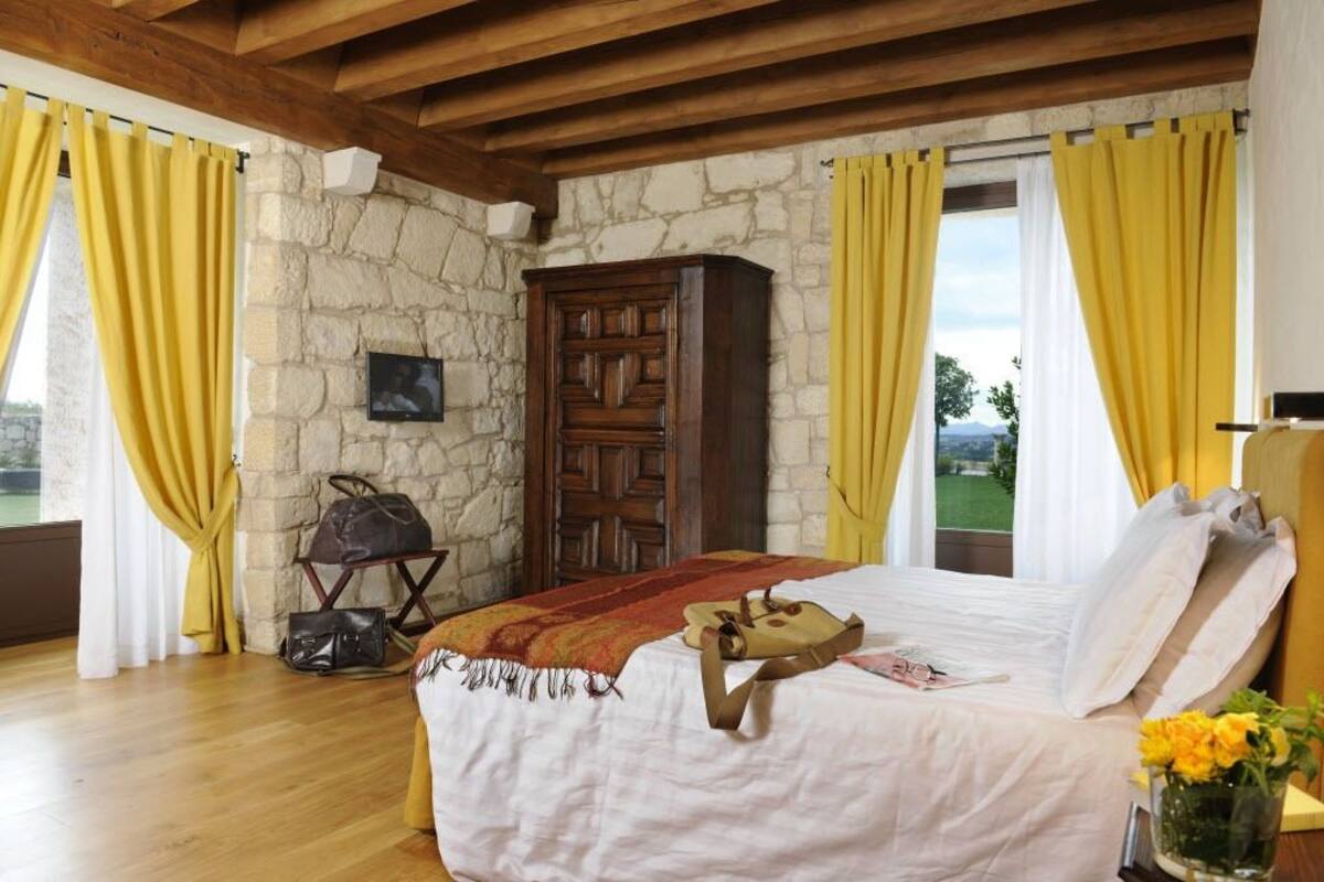 A bedroom with a stone wall, wooden ceiling beams, a bed with white linens, a wooden wardrobe, and a view through a window with yellow curtains.