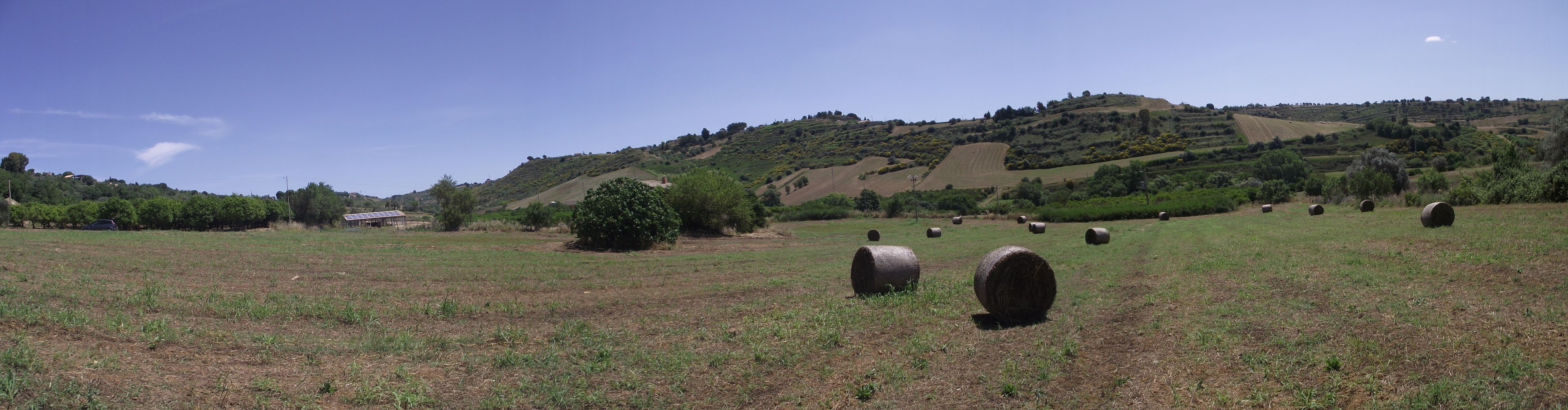 Paisaje rural con colinas ondulantes, fardos de heno y una construcción pequeña con paneles solares.