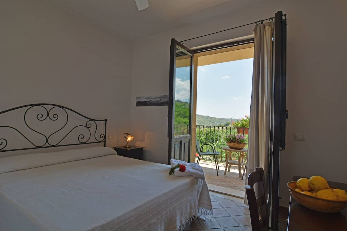 A bedroom with a bed, a nightstand, a window with curtains, a balcony with a table and chairs, and a view of greenery.