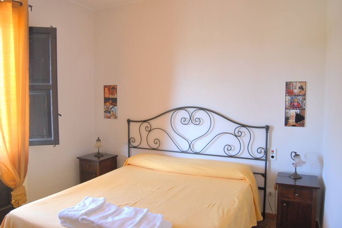 A bedroom with a metal bed frame, a yellow bedspread, a bedside table with a lamp, and a wall-mounted picture.