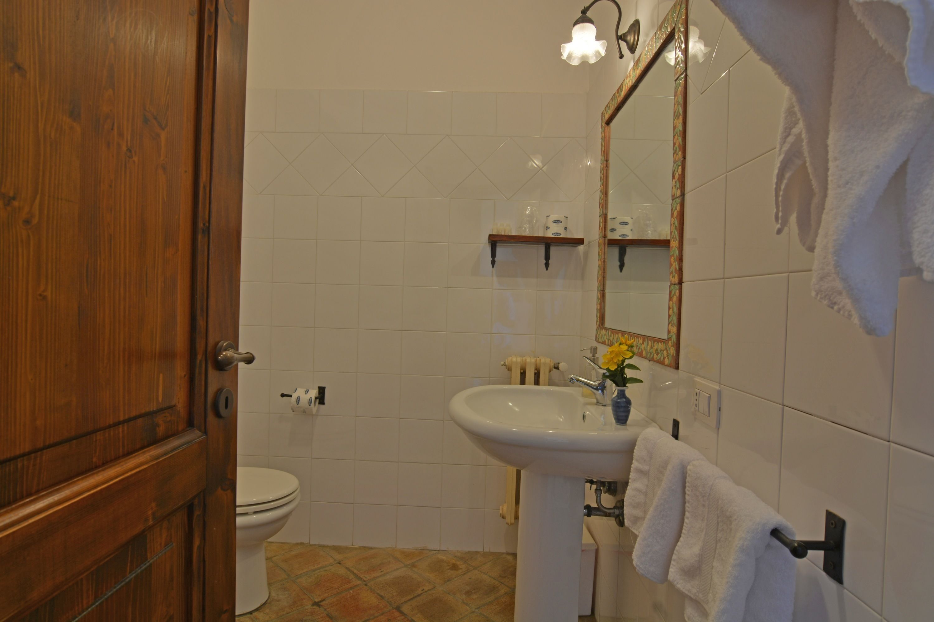 A bathroom with a wooden door, white tiles, a sink, a mirror, and a towel rack.