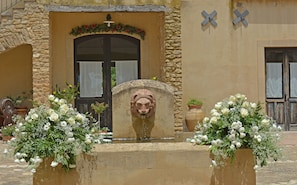 Ein Steinhaus mit einem zentralen Eingang, flankiert von je einem Blumengesteck, und einer Löwenkopf-Fontäne.