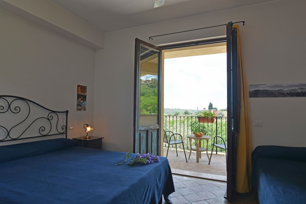 A bedroom with a bed, a nightstand, and a window leading to a balcony with a chair and table.