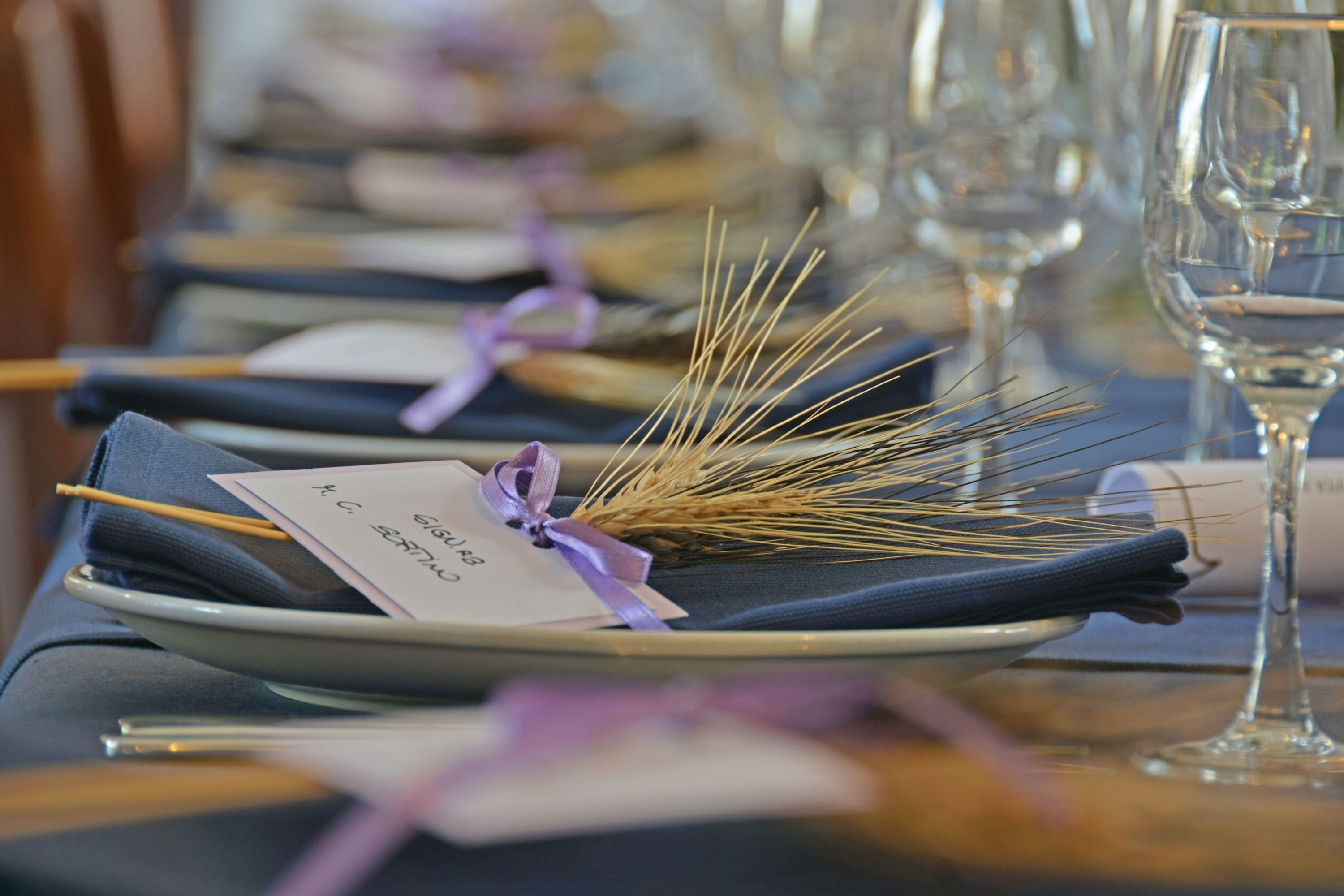 Una mesa dispuesta con platos, cubiertos y copas de vino, decorada con espigas de trigo atadas con un lazo.