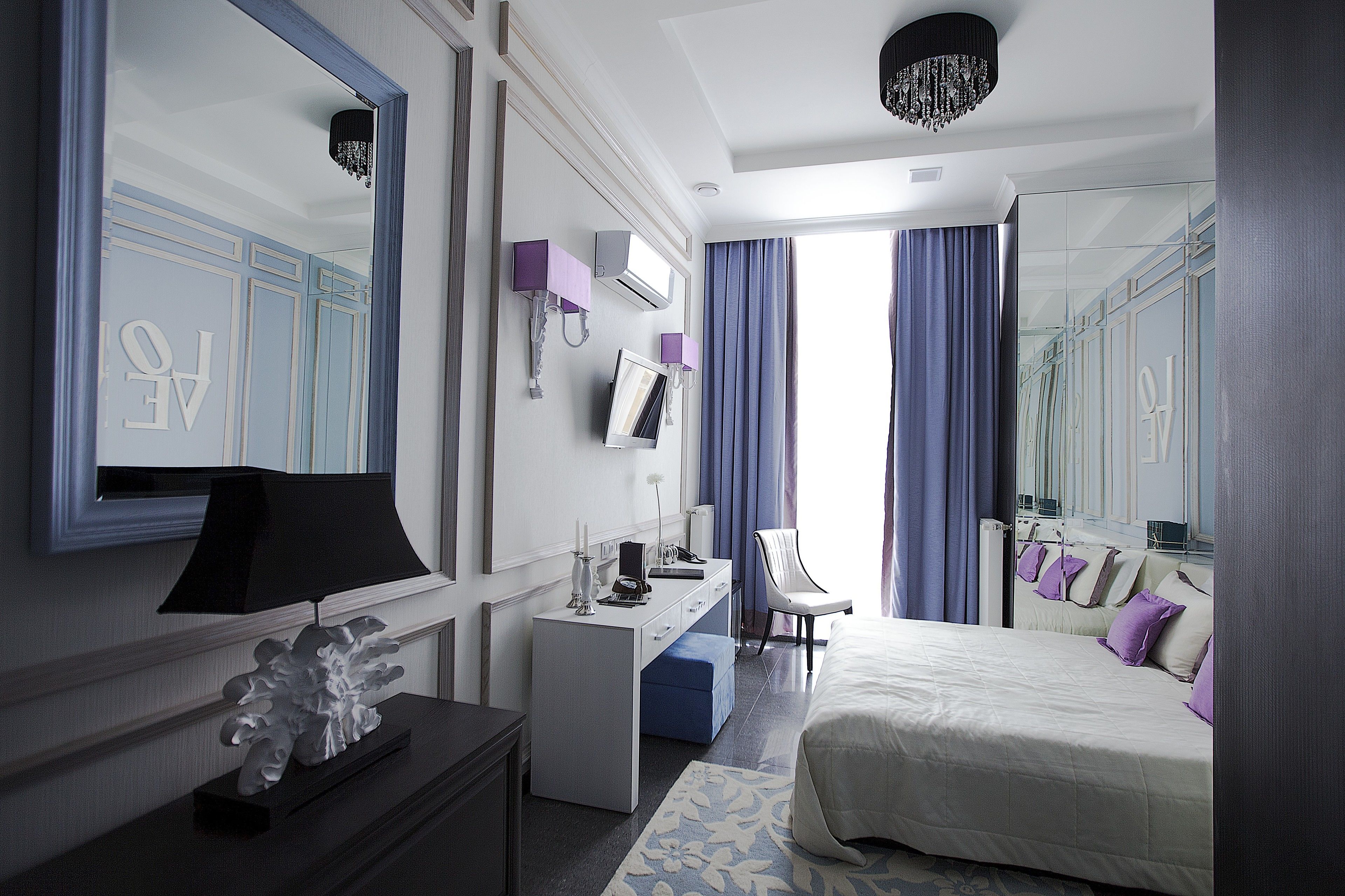 A modern hotel room with a large bed, a desk with a computer, a chair, and a large mirror.