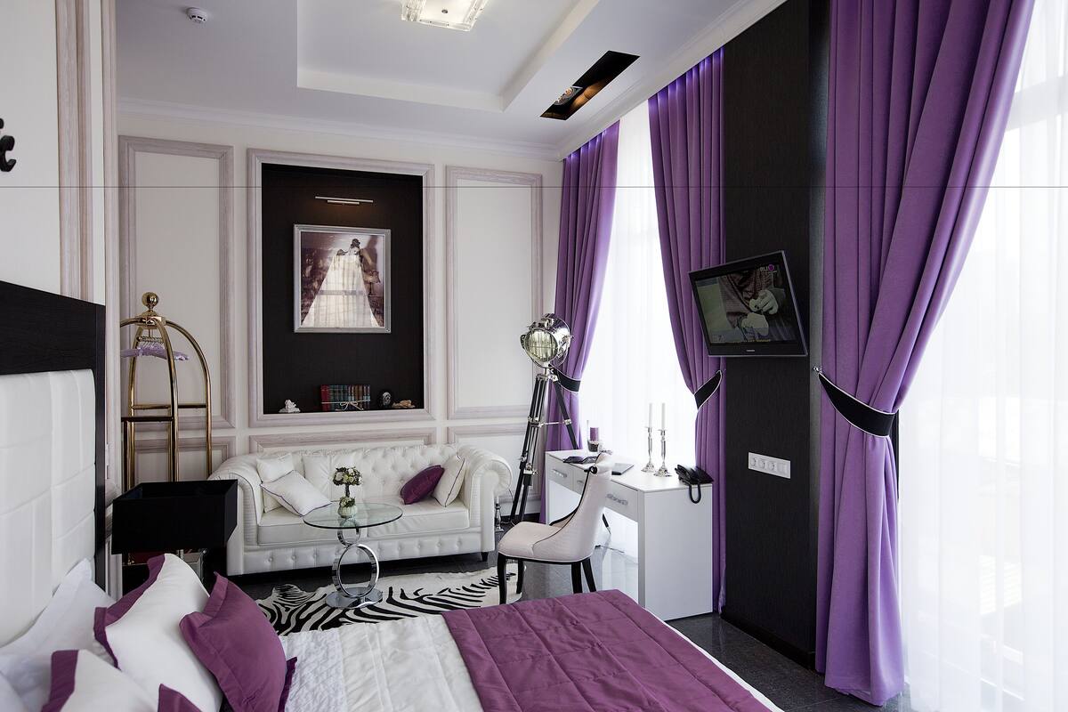 Habitación de hotel con cama, escritorio, silla, sofá, televisor y cortinas moradas.