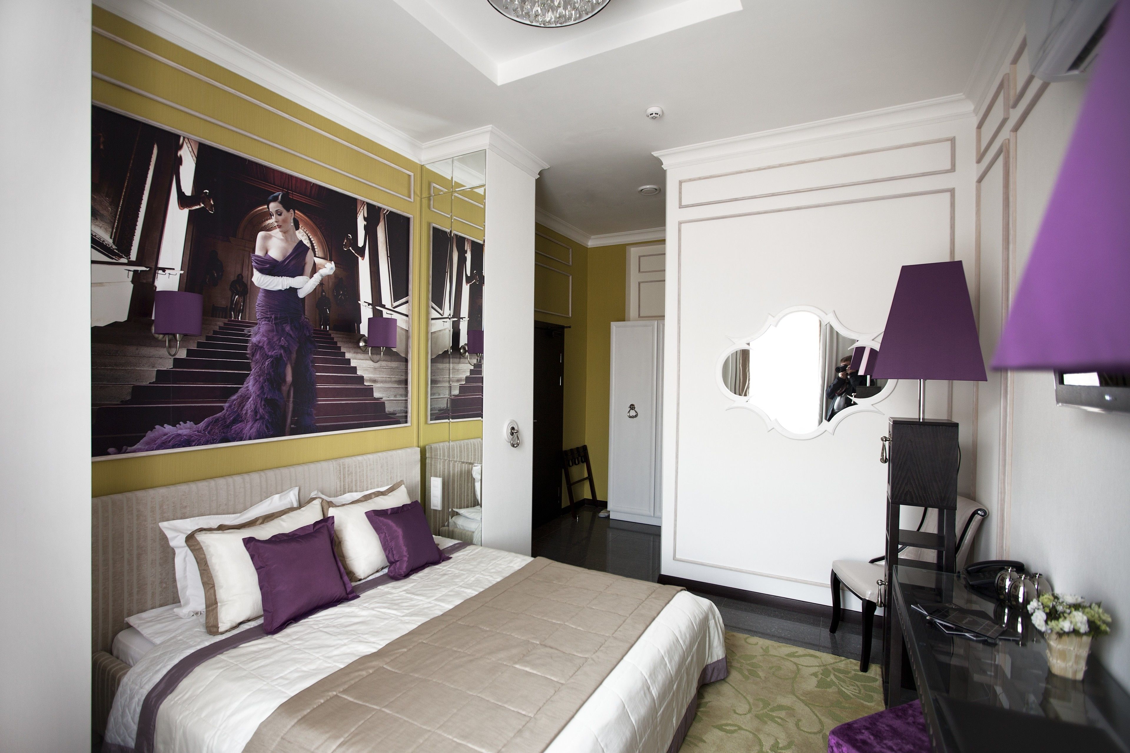 A modern hotel room with a large bed, a mirror, a desk, and a view of a staircase.