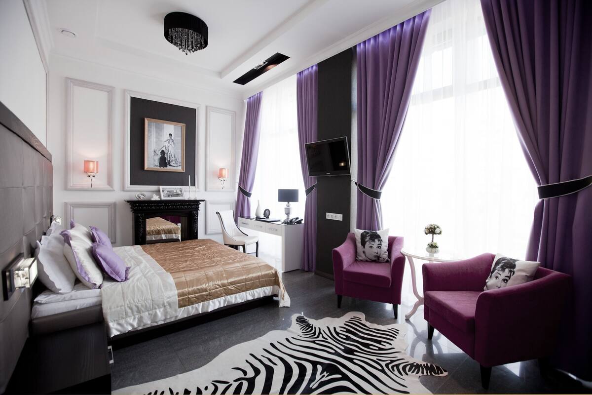 Una habitación de hotel moderna con una cama, un escritorio, una silla, un televisor y cortinas moradas.