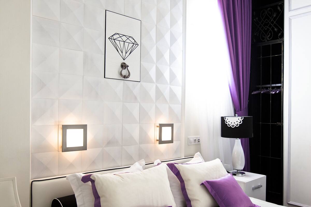 Una habitación de hotel moderna con un cabecero de patrón geométrico, una cama con ropa de cama blanca y morada, dos mesitas de noche con lámparas, y un ventanal con cortinas moradas.