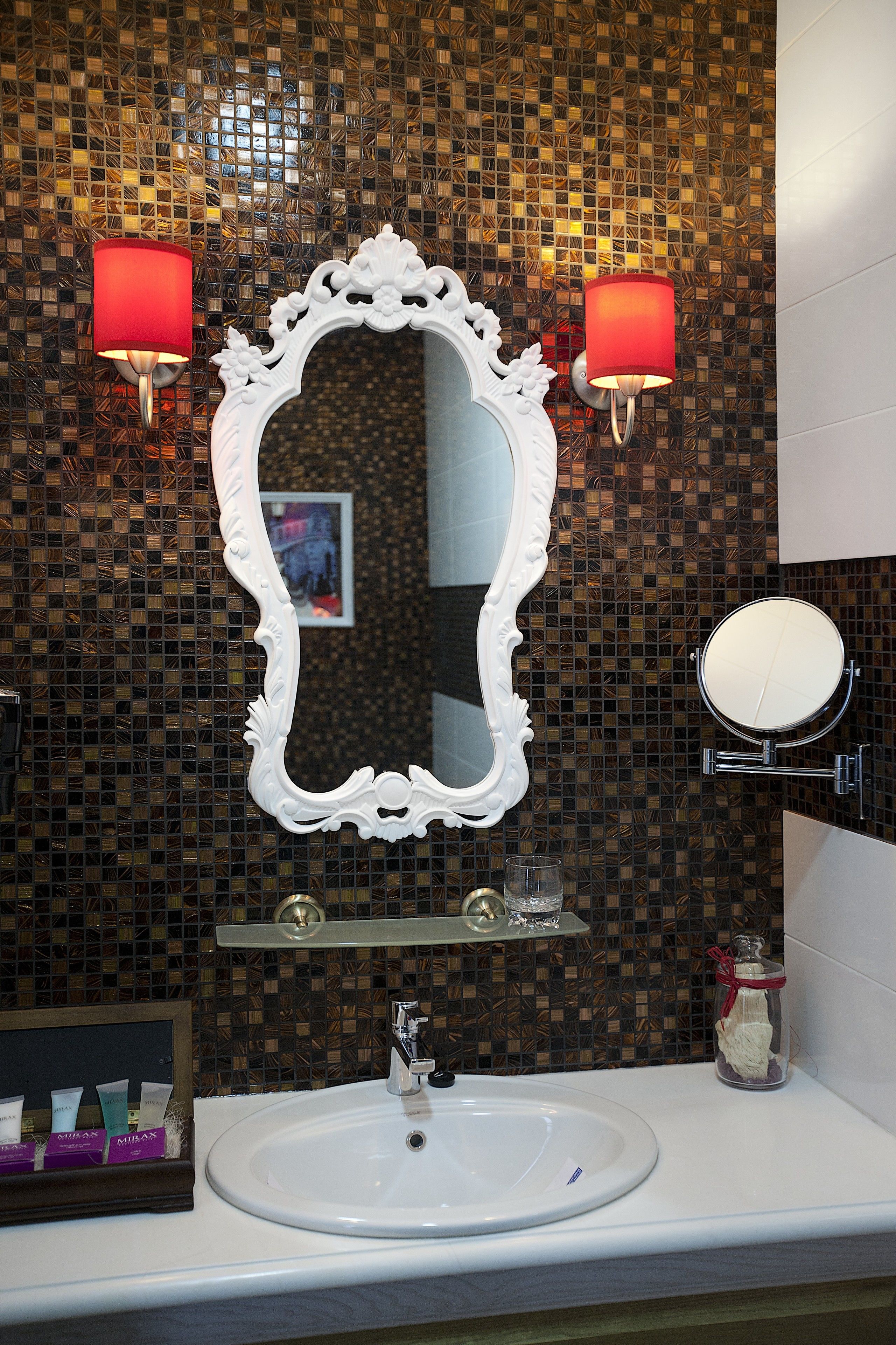 Ein Badezimmer mit einem weißen Waschbecken, einem großen, verzierten Spiegel und zwei Wandleuchten mit roten Lampen.