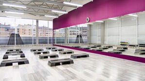 Amplio espacio interior con varias estaciones de ejercicio sobre un piso de tonos claros, grandes ventanales que ofrecen vistas de edificios exteriores y una pared de acento morada.