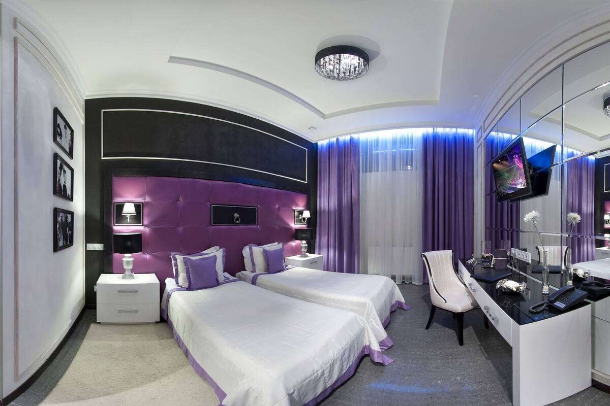 Una habitación de hotel moderna con techo curvo, dos camas con ropa de cama morada, una tocador con espejo y un televisor.
