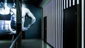 Un pasillo con un muro espejado que refleja una figura musculosa, una barandilla de metal y una pared con tiras de luz verticales.