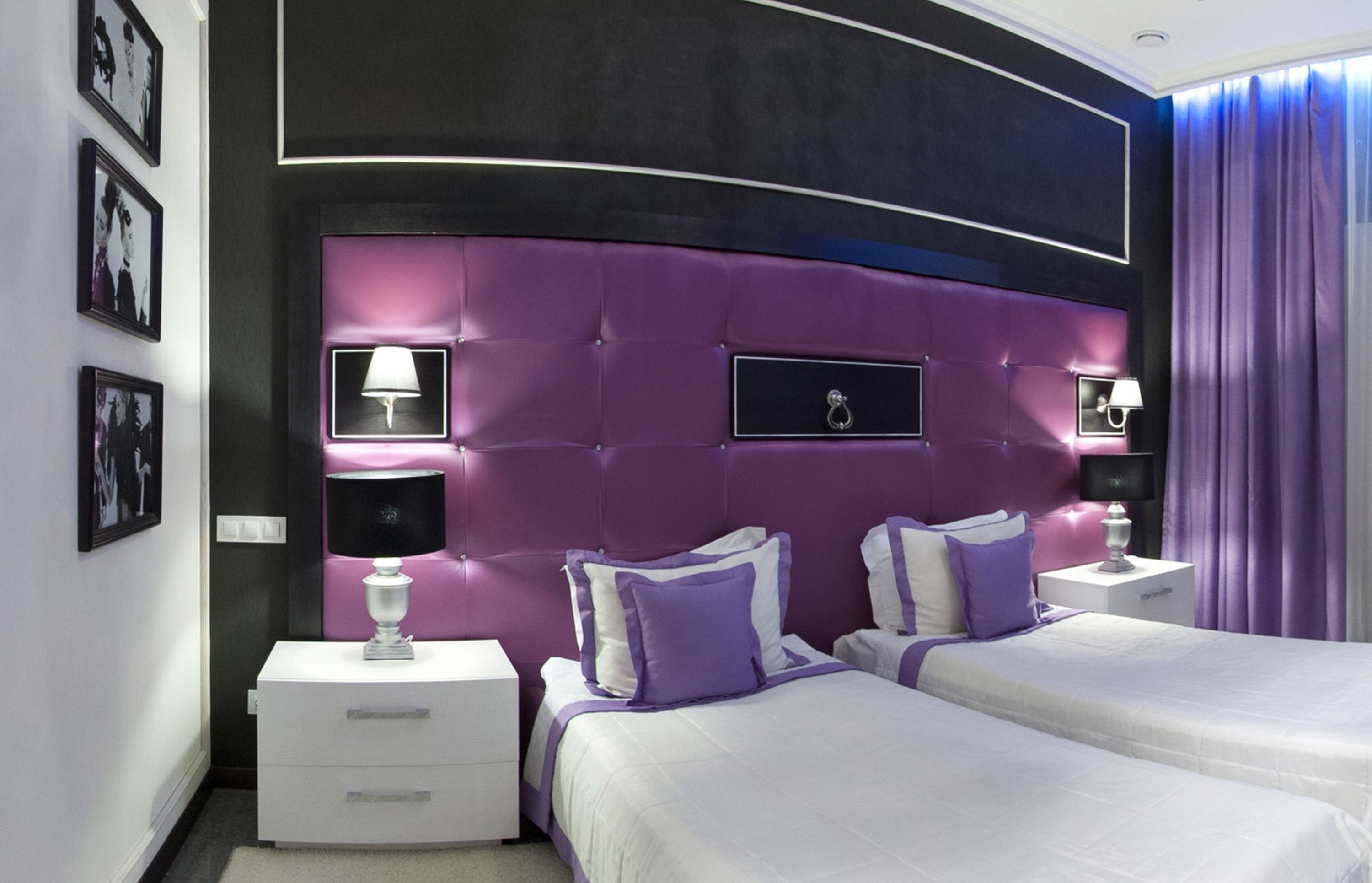 A modern bedroom with a curved headboard, two beds, white nightstands, and purple bedding.