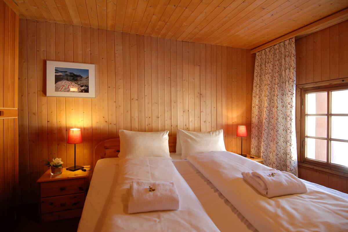 A wooden-paneled room with two beds, each with a pillow and a folded towel. There is a nightstand with a lamp and a framed picture on the wall.