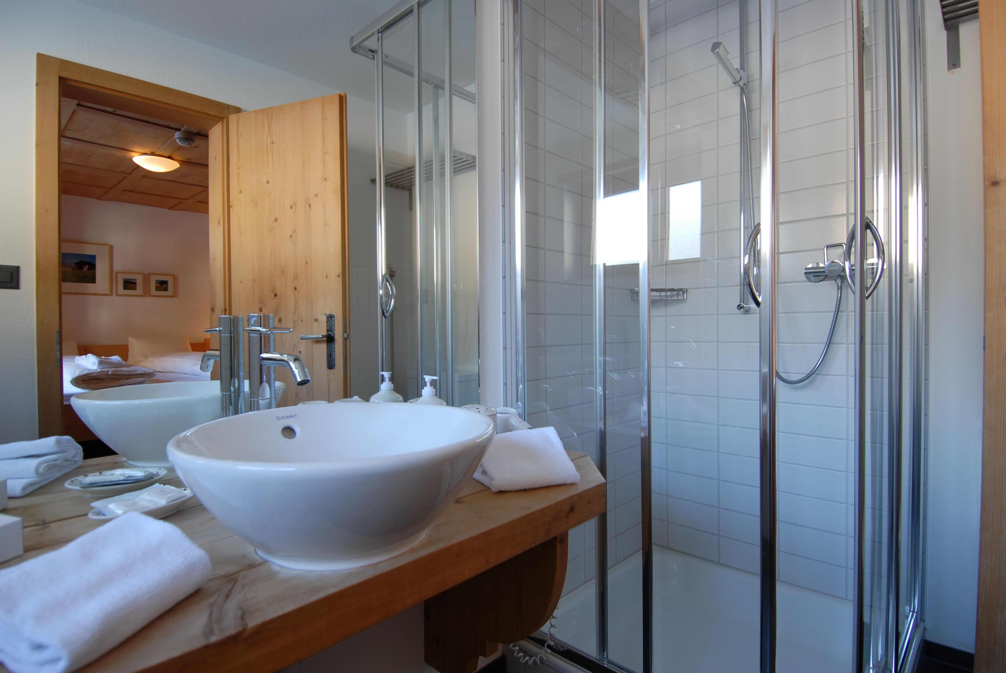 Une salle de bain moderne avec un grand lavabo blanc, un meuble-lavabo en bois, une douche à parois de verre et une porte en bois donnant sur une autre pièce.