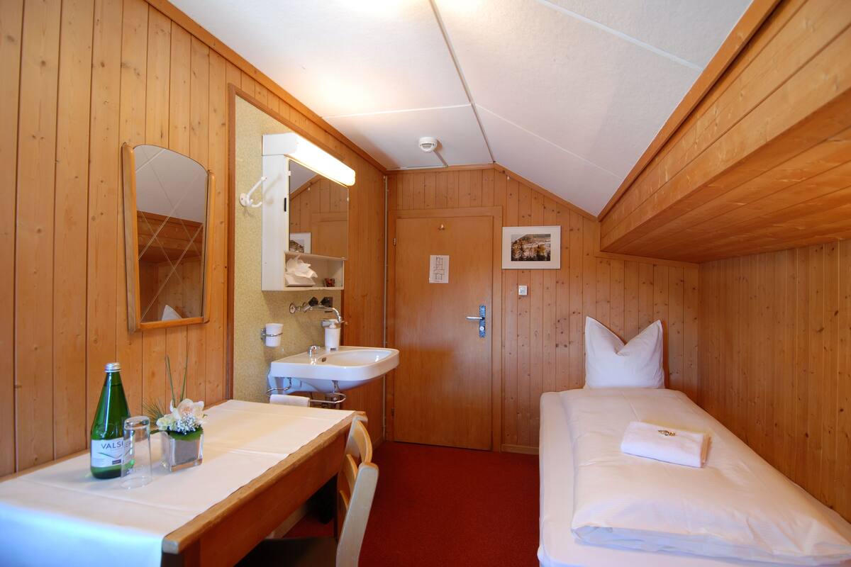 A wooden cabin room with a wooden bench, a small table with a bottle and glasses, a sink, and a bed with pillows.