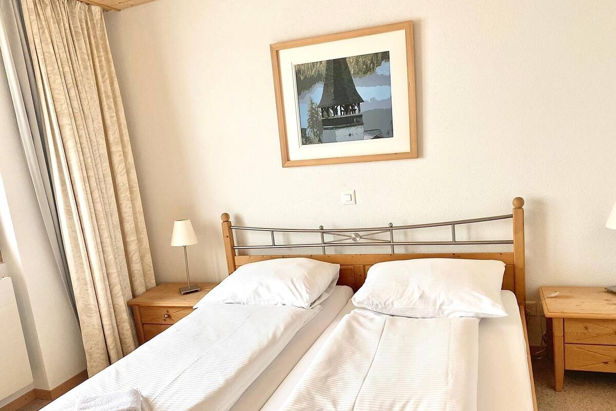 A double bed with two pillows and a folded towel, a bedside table with a lamp, and a framed picture on the wall.
