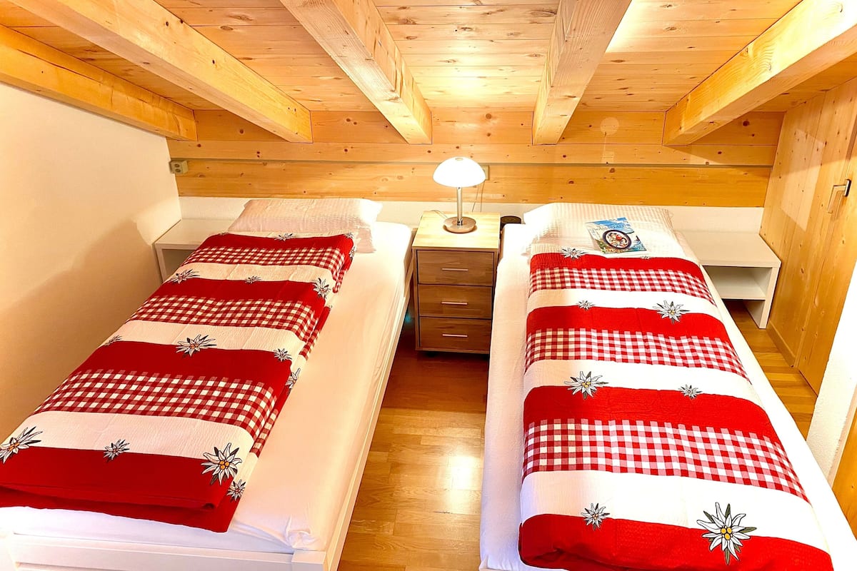A bedroom with two single beds, wooden ceiling beams, a nightstand with a lamp, and a small drawer.