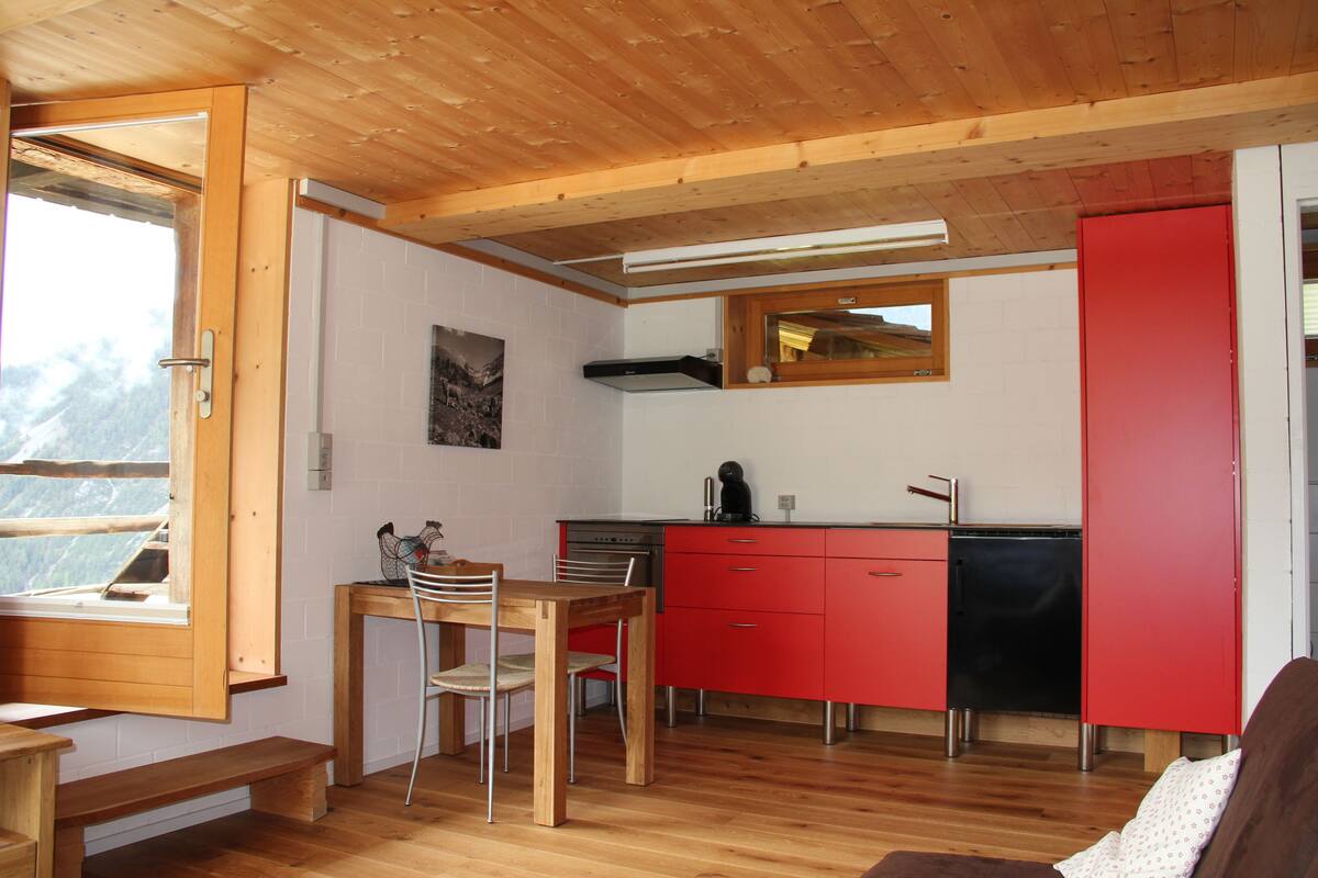 A compact kitchen with a wooden table and chairs, a red cabinet, and a window with a view of mountains.