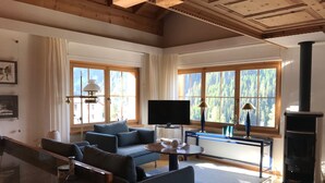 A spacious living room with a sofa, TV, and fireplace. Large windows offer views of a forested area.