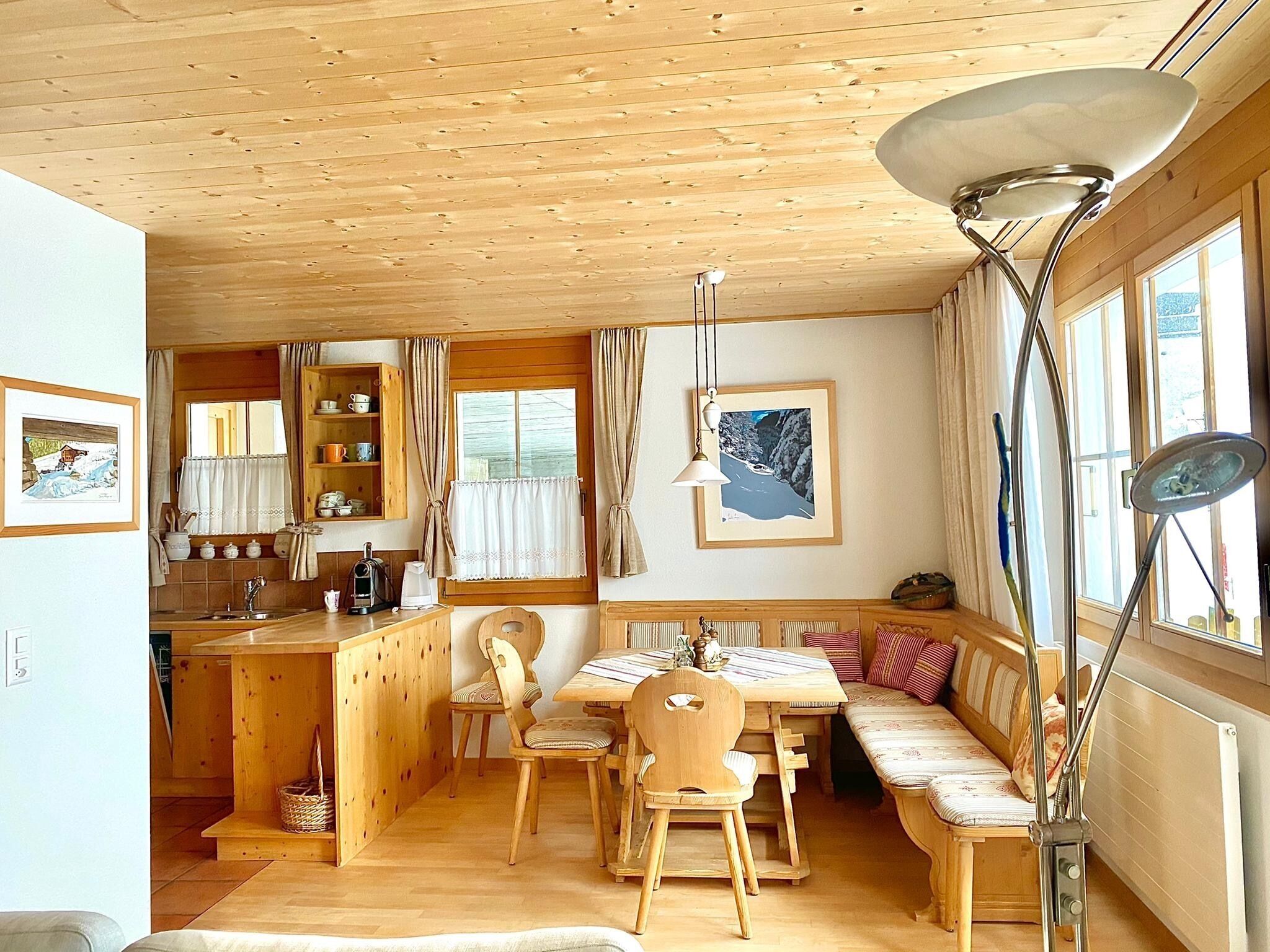 A cozy cabin interior with a wooden dining table, chairs, a bench, a kitchen area, and a large window with curtains.
