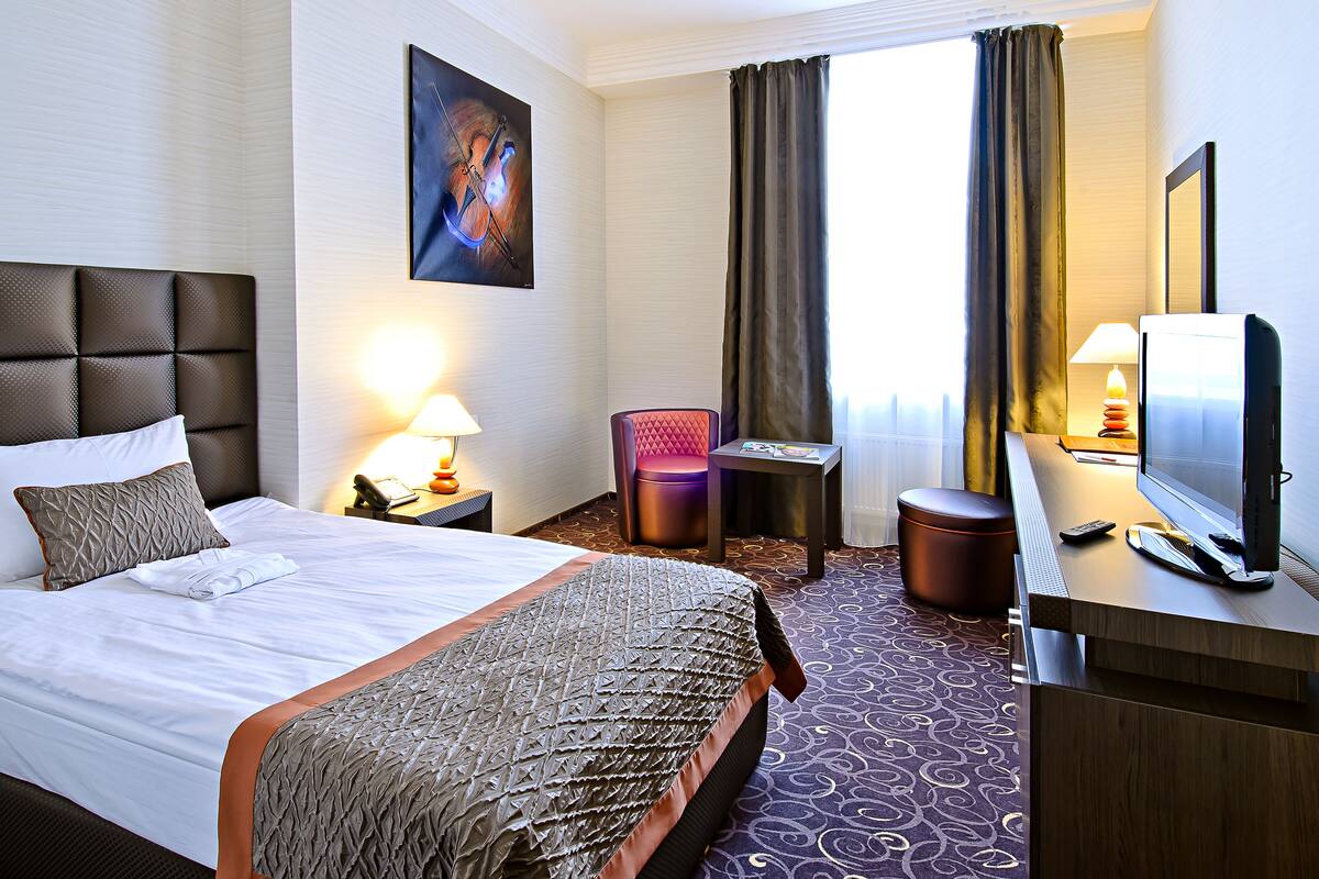 Ein Hotelzimmer mit einem großen Bett, einem Schreibtisch mit Fernseher, einem Sessel, einer Lampe und einem Bild an der Wand.