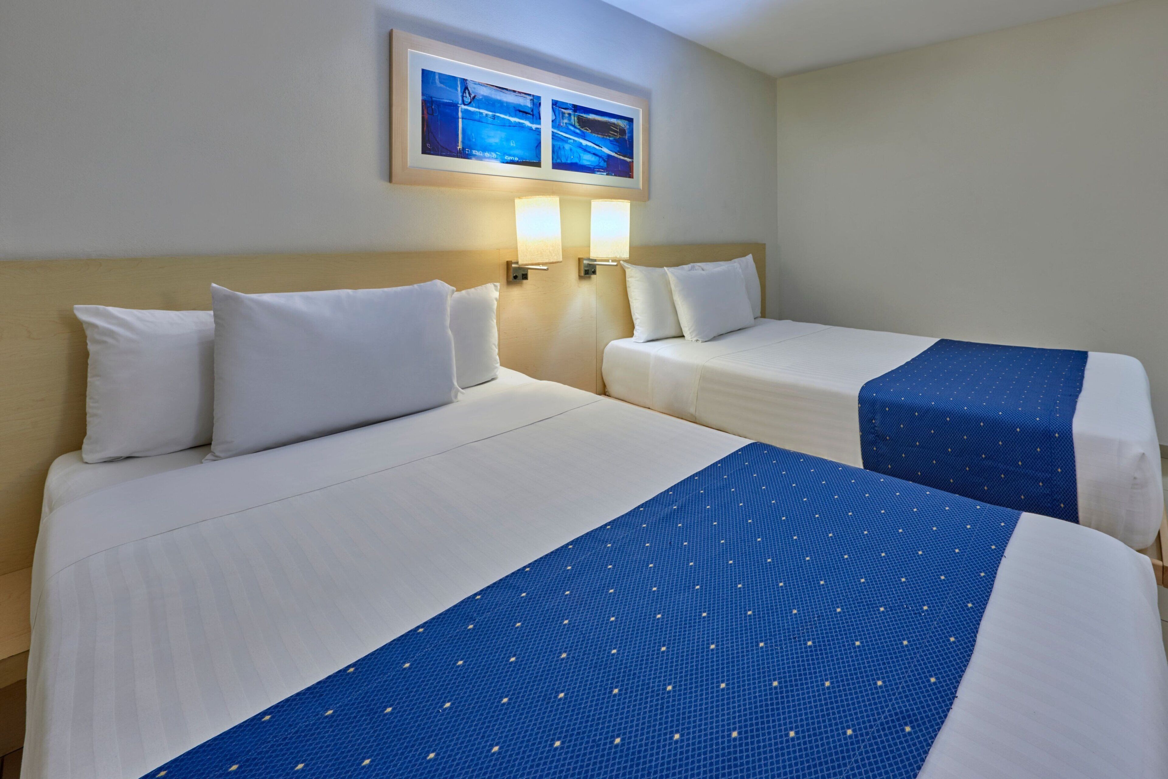 A hotel room with two beds, each with white bedding and a blue accent. There are wall-mounted lights and a framed picture above the beds.