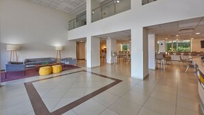 Eine großzügige Lobby mit hoher Decke, eine Sitzecke mit einer Couch und Sesseln sowie ein Essbereich mit Tischen und Stühlen.