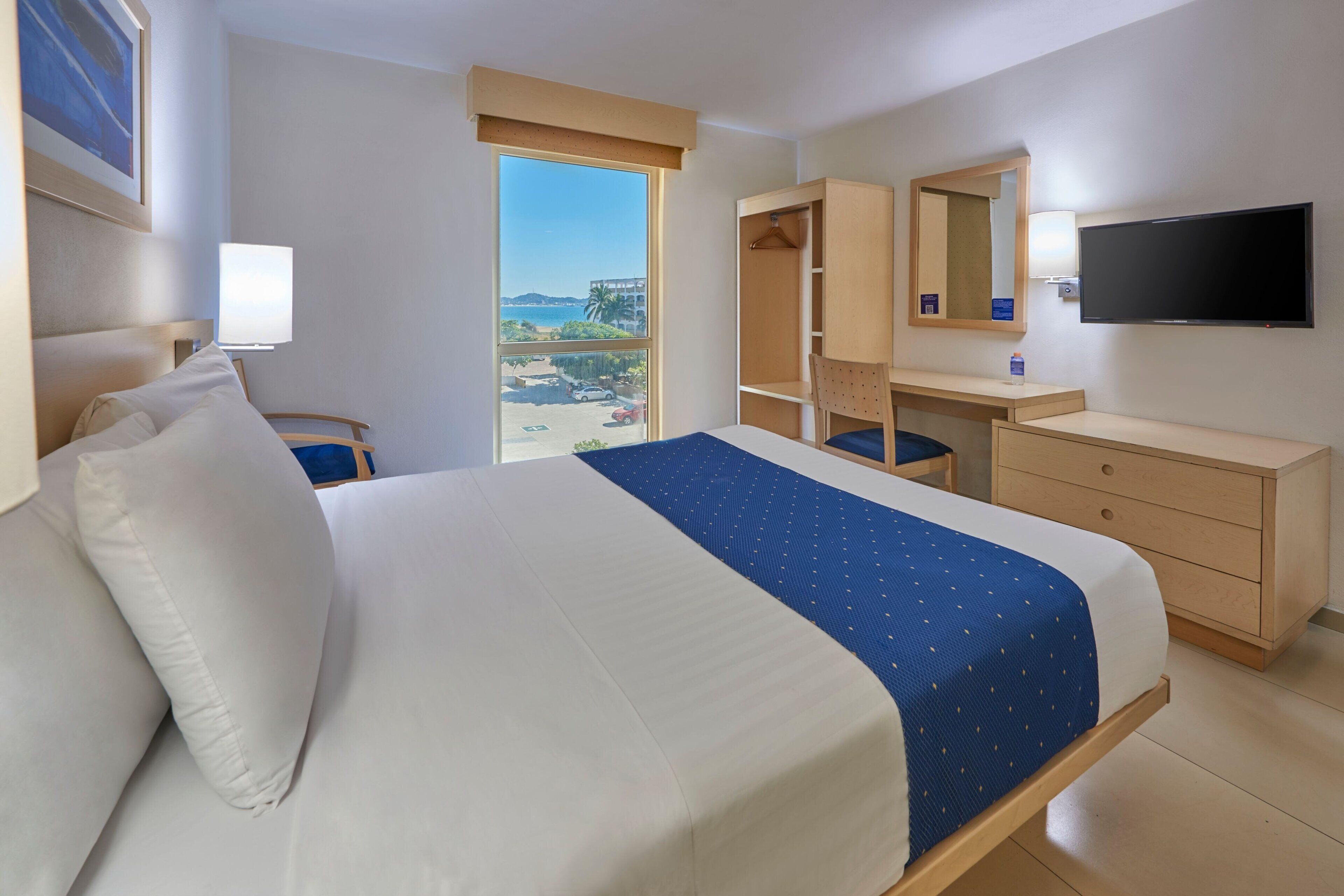 A hotel room with a large bed, a desk, a chair, a wardrobe, a TV, and a window with a view of the sea.