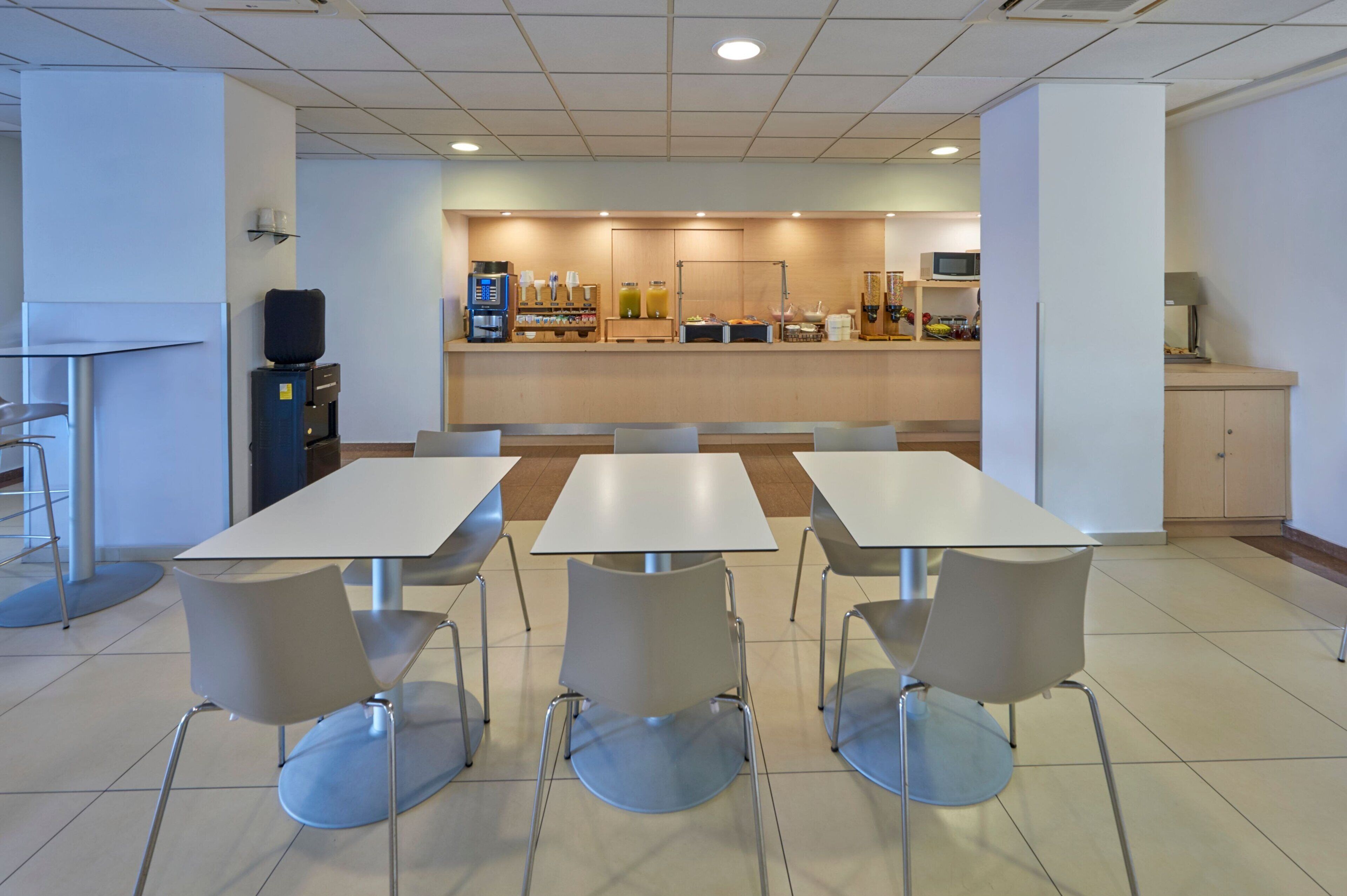 Una cafetería moderna con mesas y sillas blancas, una estación de café y un microondas.
