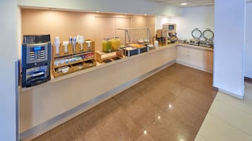 Un área de desayuno autoservicio con una máquina de café, dispensadores de bebidas y diversas opciones de comida.