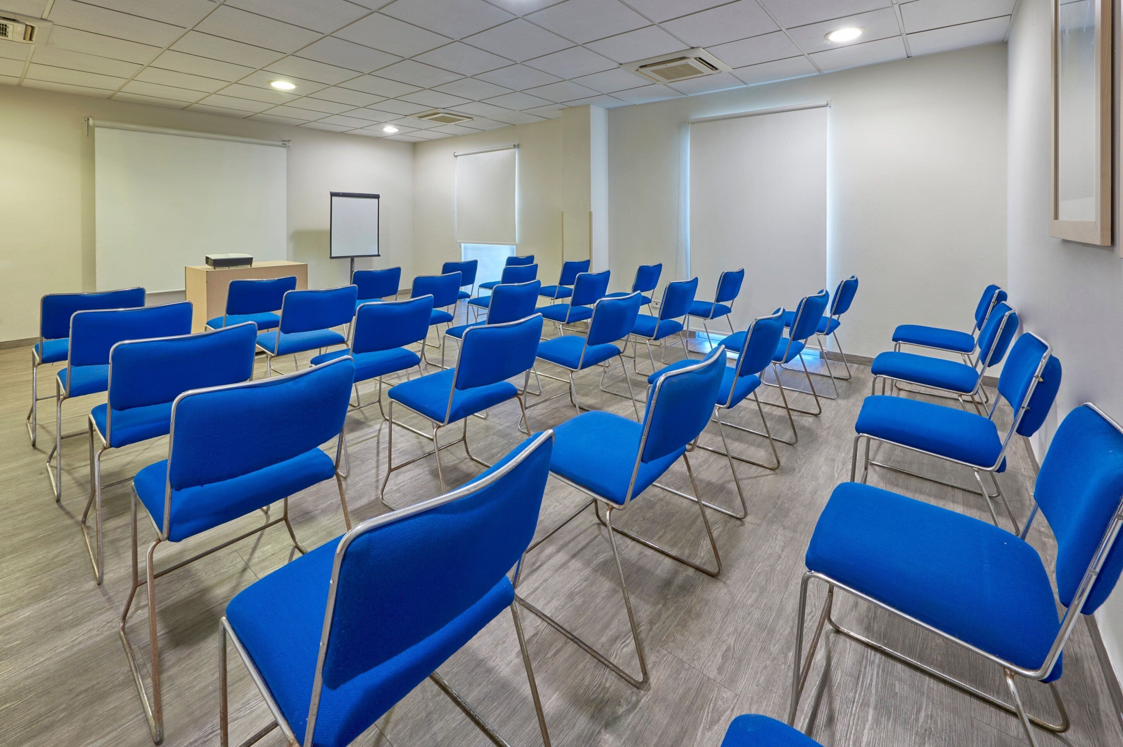 Ein Konferenzraum mit blauen Stühlen, einem Rednerpult, einer Tafel und einer Projektionsleinwand.