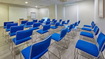 Une salle de conférence avec des rangées de chaises bleues, un pupitre, un tableau blanc et un écran de projection.