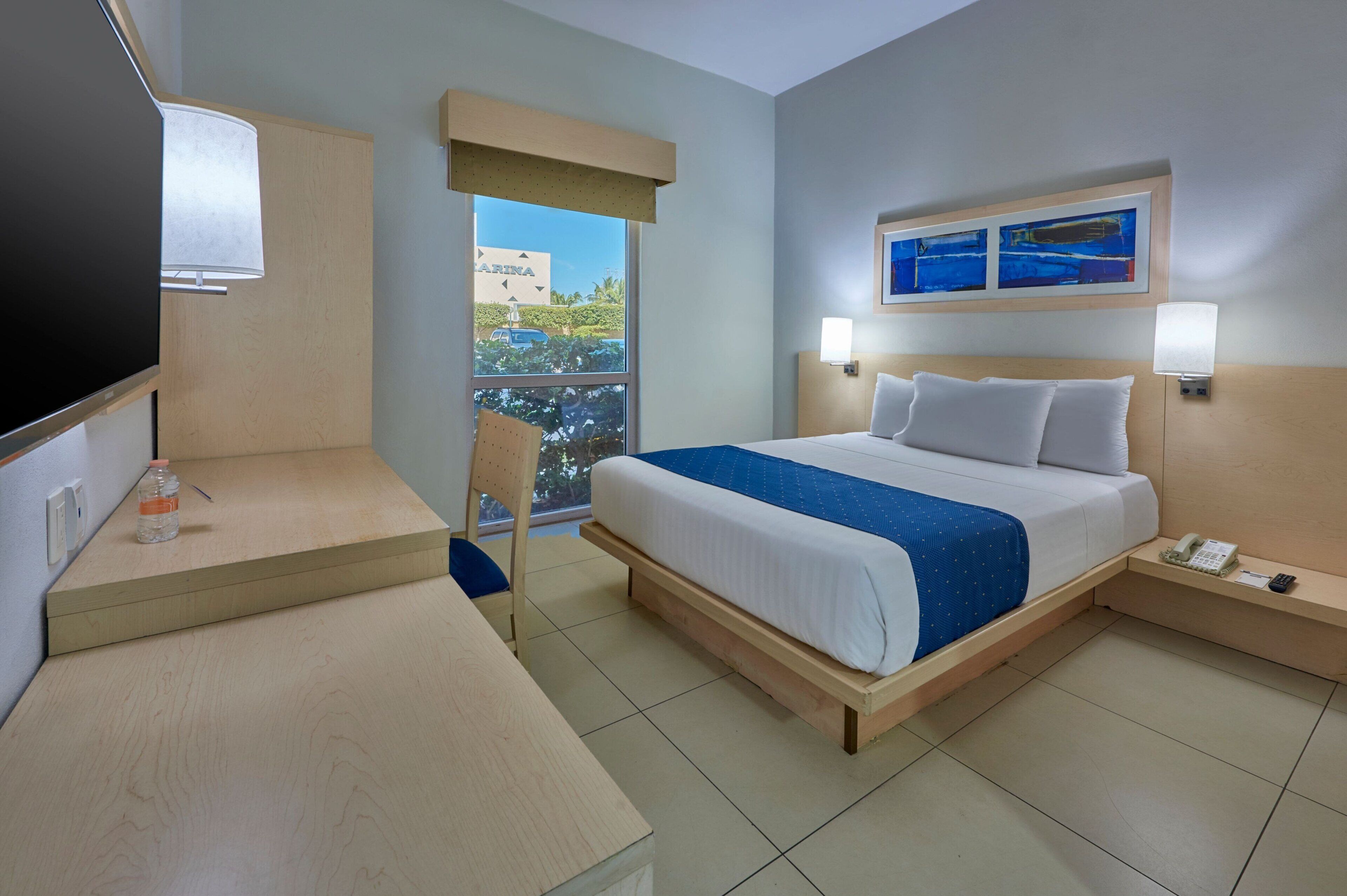 Habitación de hotel con cama, escritorio, silla, televisión, teléfono y ventana con vistas a un edificio y vegetación.