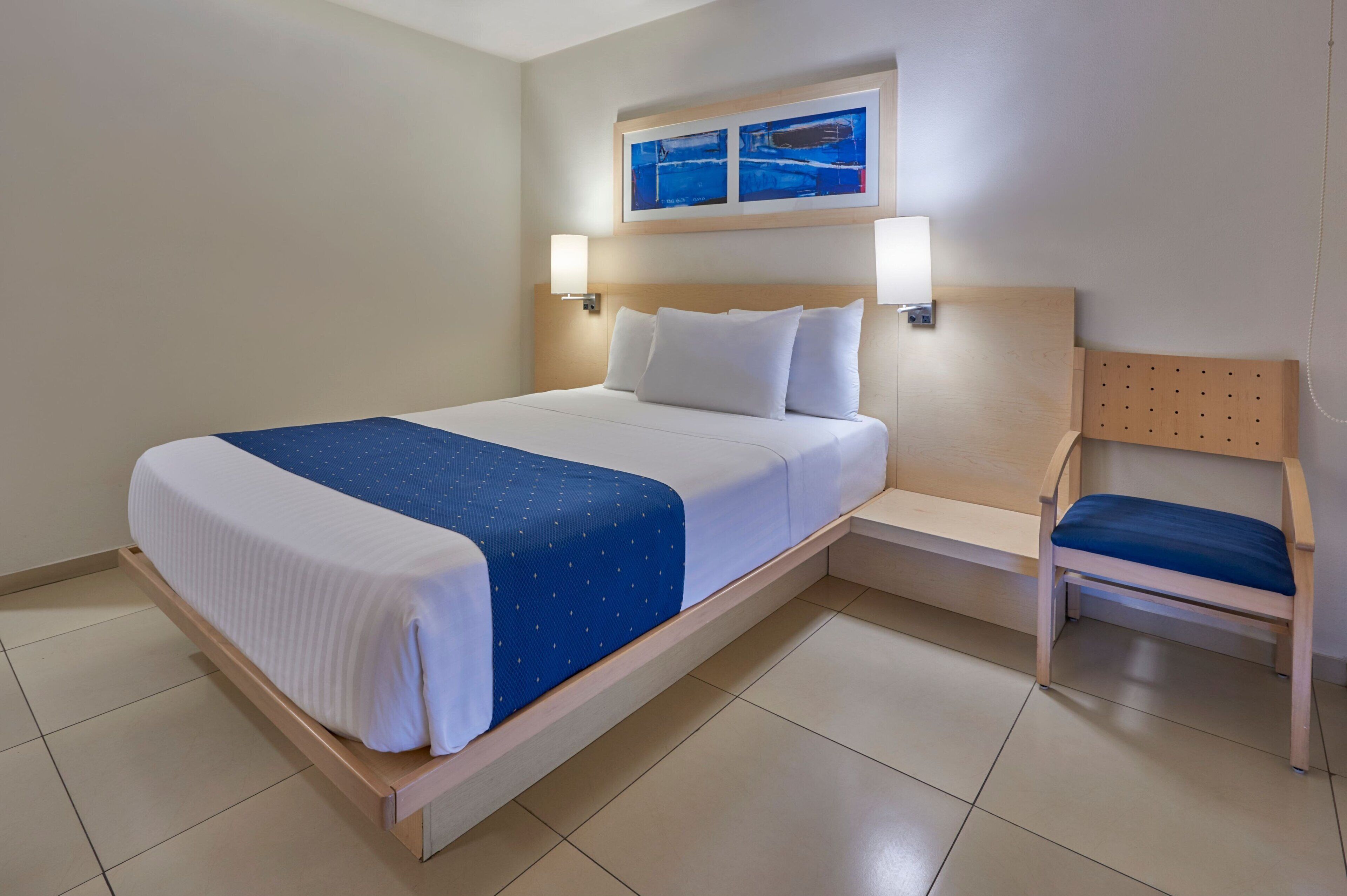 A hotel room with a bed, a chair, and two wall-mounted lamps.