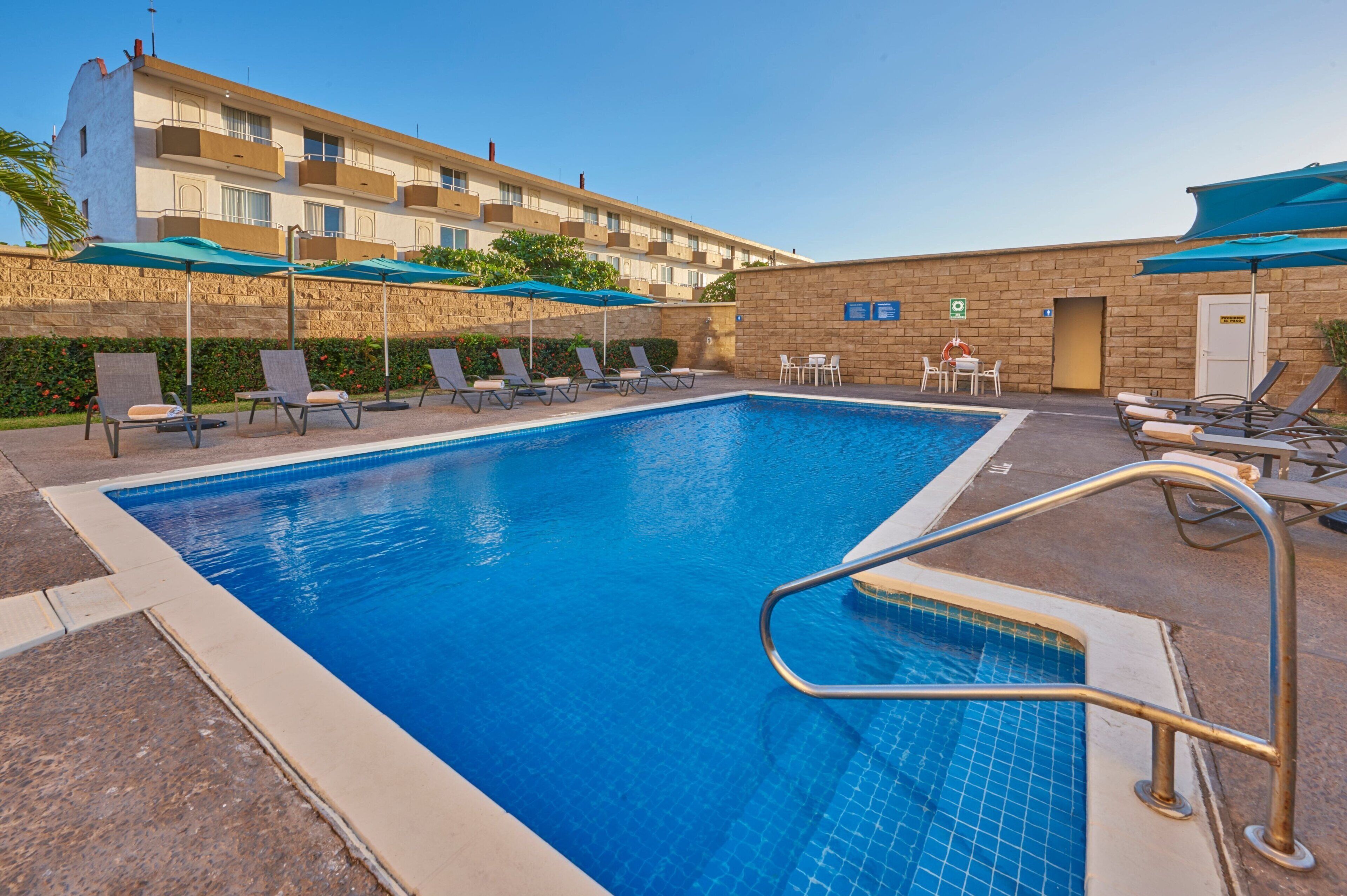 A swimming pool with lounge chairs and umbrellas, surrounded by a stone wall and a building with balconies.