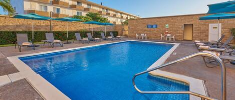 A swimming pool with lounge chairs and umbrellas, surrounded by a stone wall and a building with balconies.