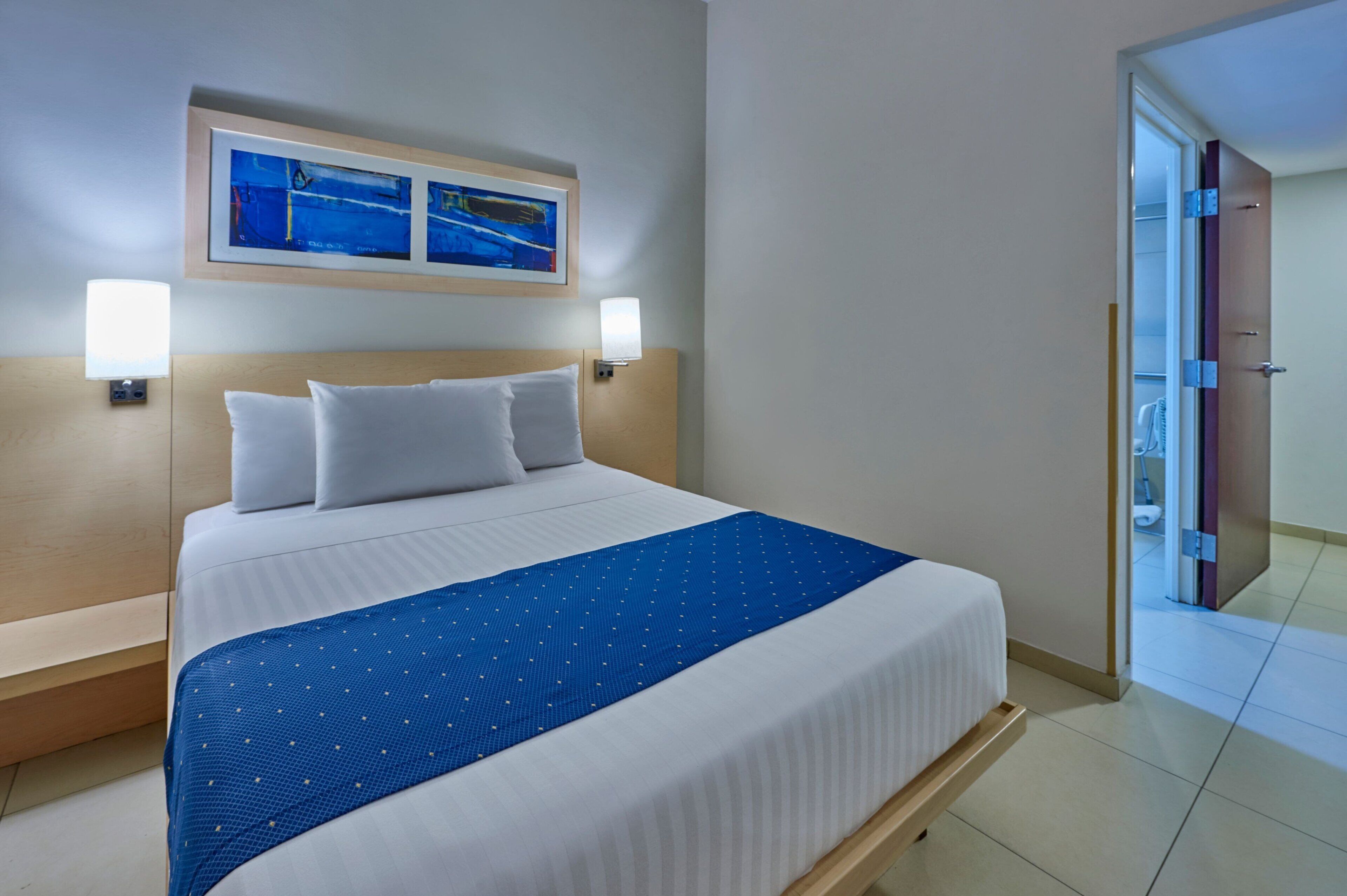 A hotel room with a large bed, two wall-mounted lamps, a blue bedspread, and a framed picture above the bed.