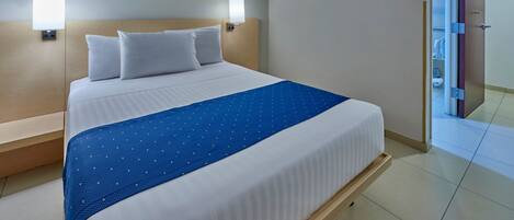 A hotel room with a large bed, two wall-mounted lamps, a blue bedspread, and a framed picture above the bed.