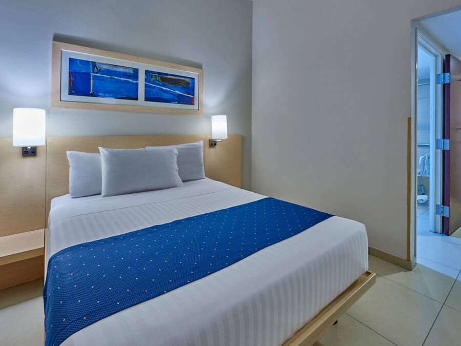A hotel room with a large bed, two wall-mounted lamps, a blue bedspread, and a framed picture above the bed.