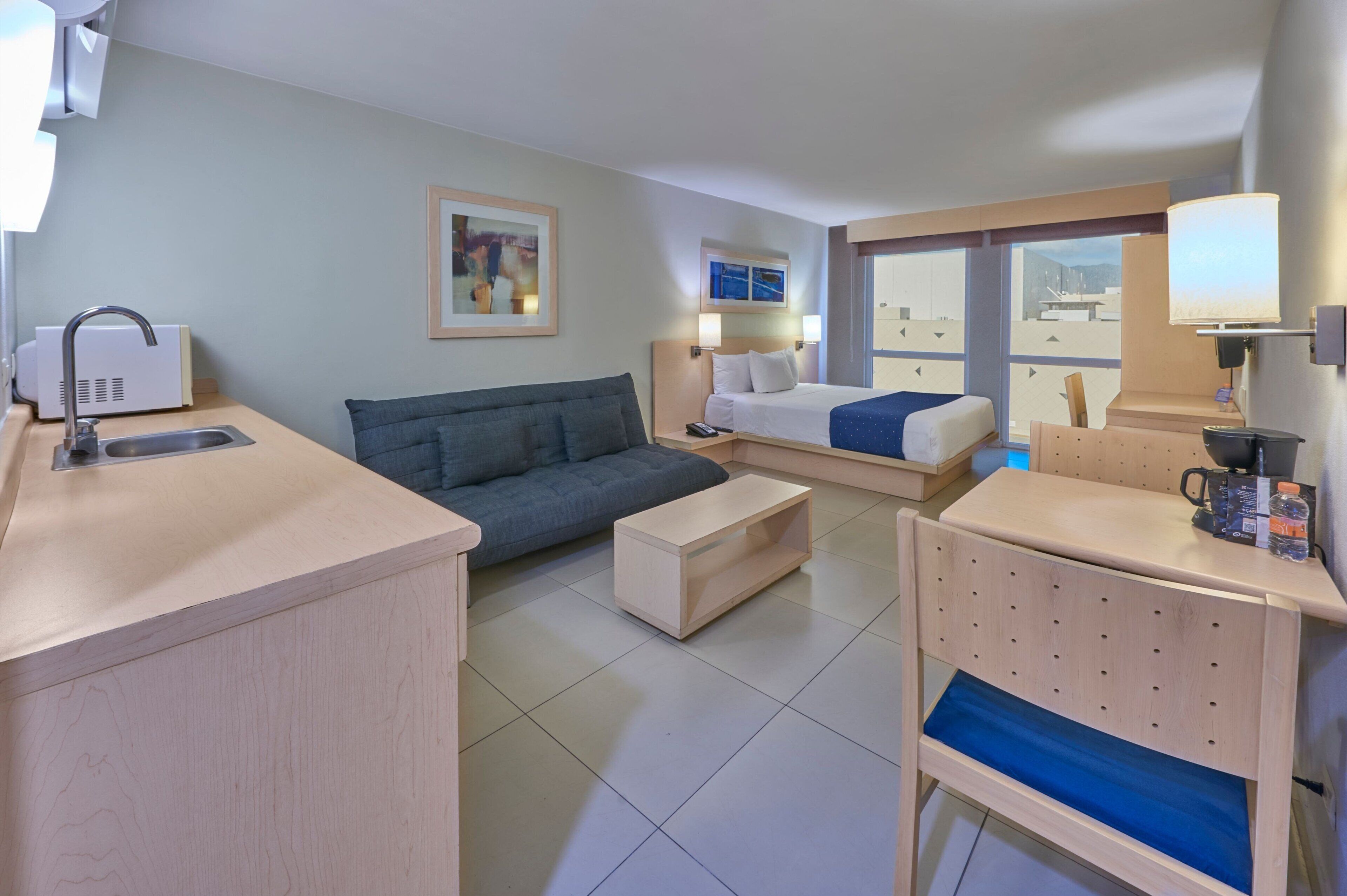 A hotel room with a kitchenette, a sofa, a bed, and a desk with a lamp.
