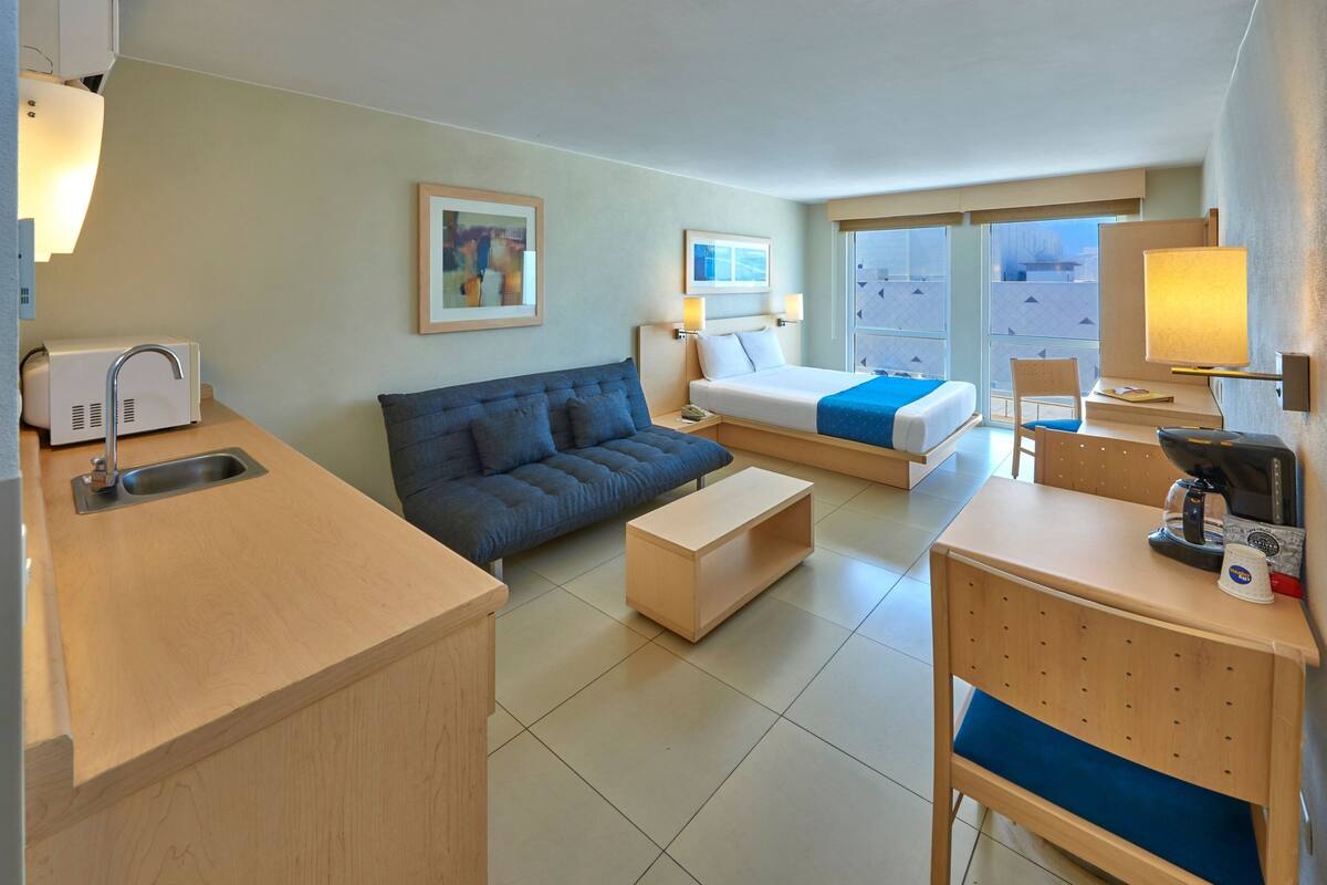 A hotel room with a kitchenette, a sofa, a bed, a desk, and a chair.