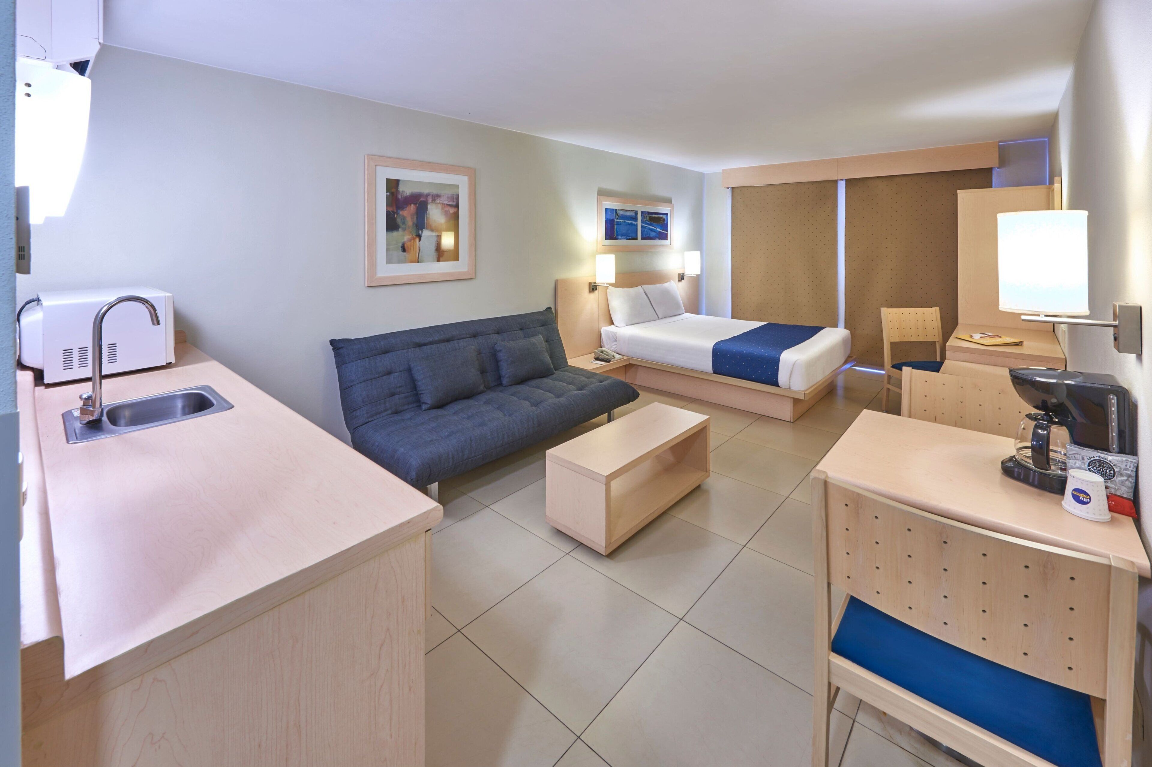 A hotel room with a kitchenette, a sofa, a bed, a desk, and a chair.